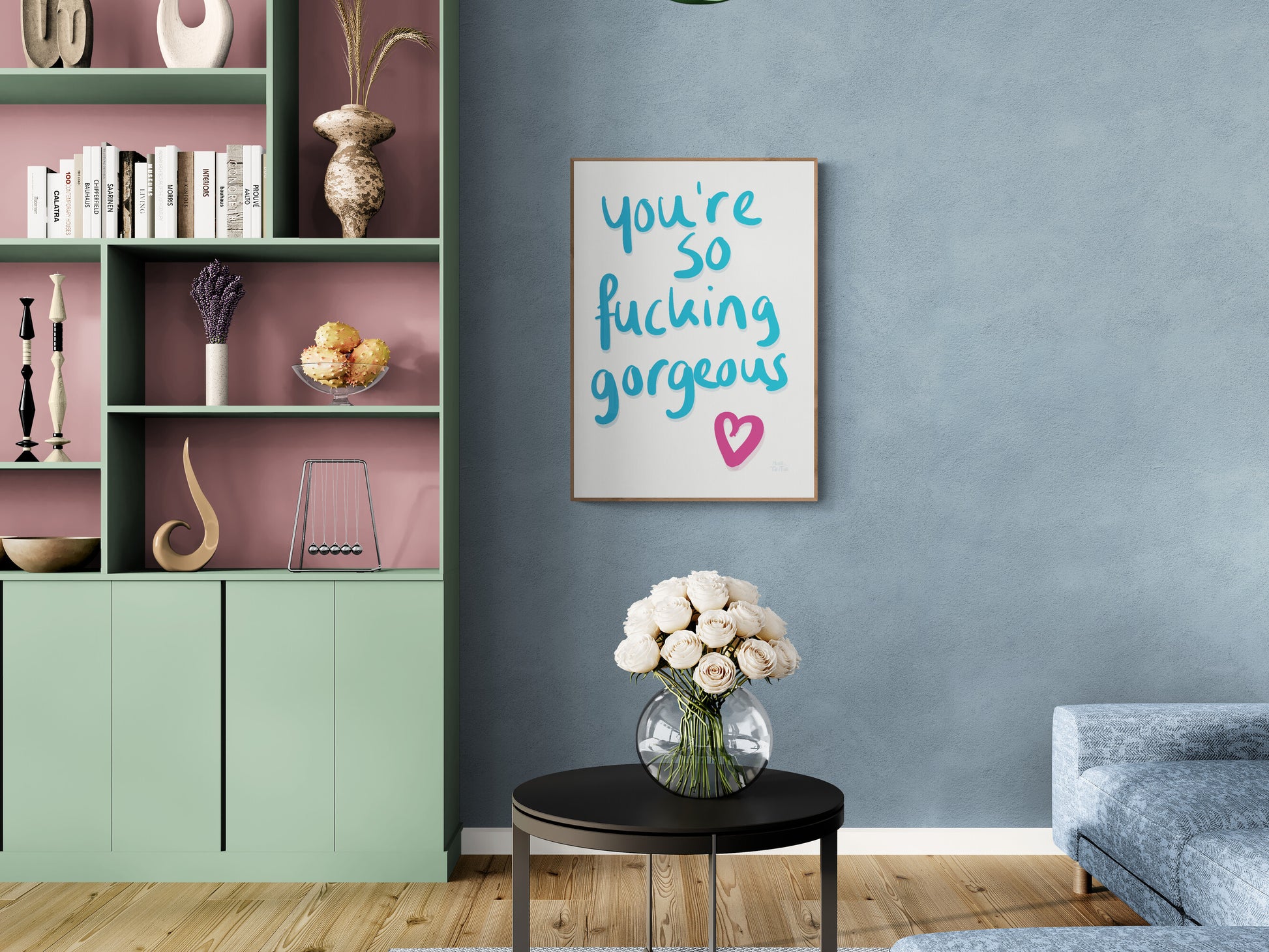 Poster with ‘You’re so fucking gorgeous’ in blue handwriting with a pink heart on a white background. Hanging on a blue wall next to a colourful cabinet in an oak frame.