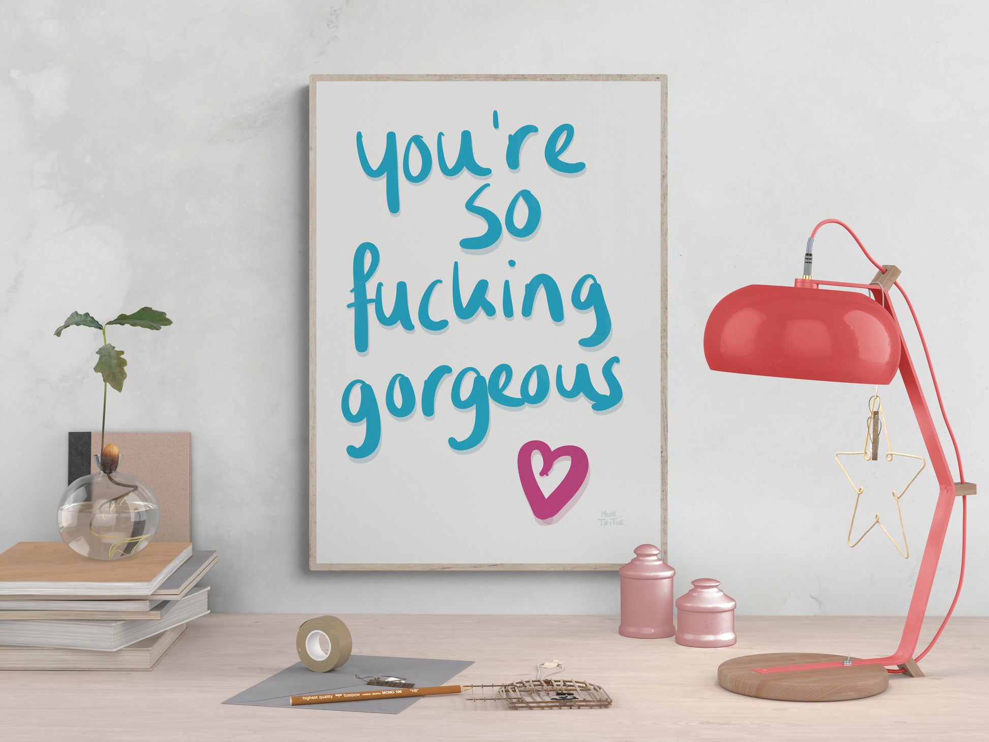 Poster with ‘You’re so fucking gorgeous’ in blue handwriting with a pink heart on a white background. Hanging above a desk in an light oak frame.