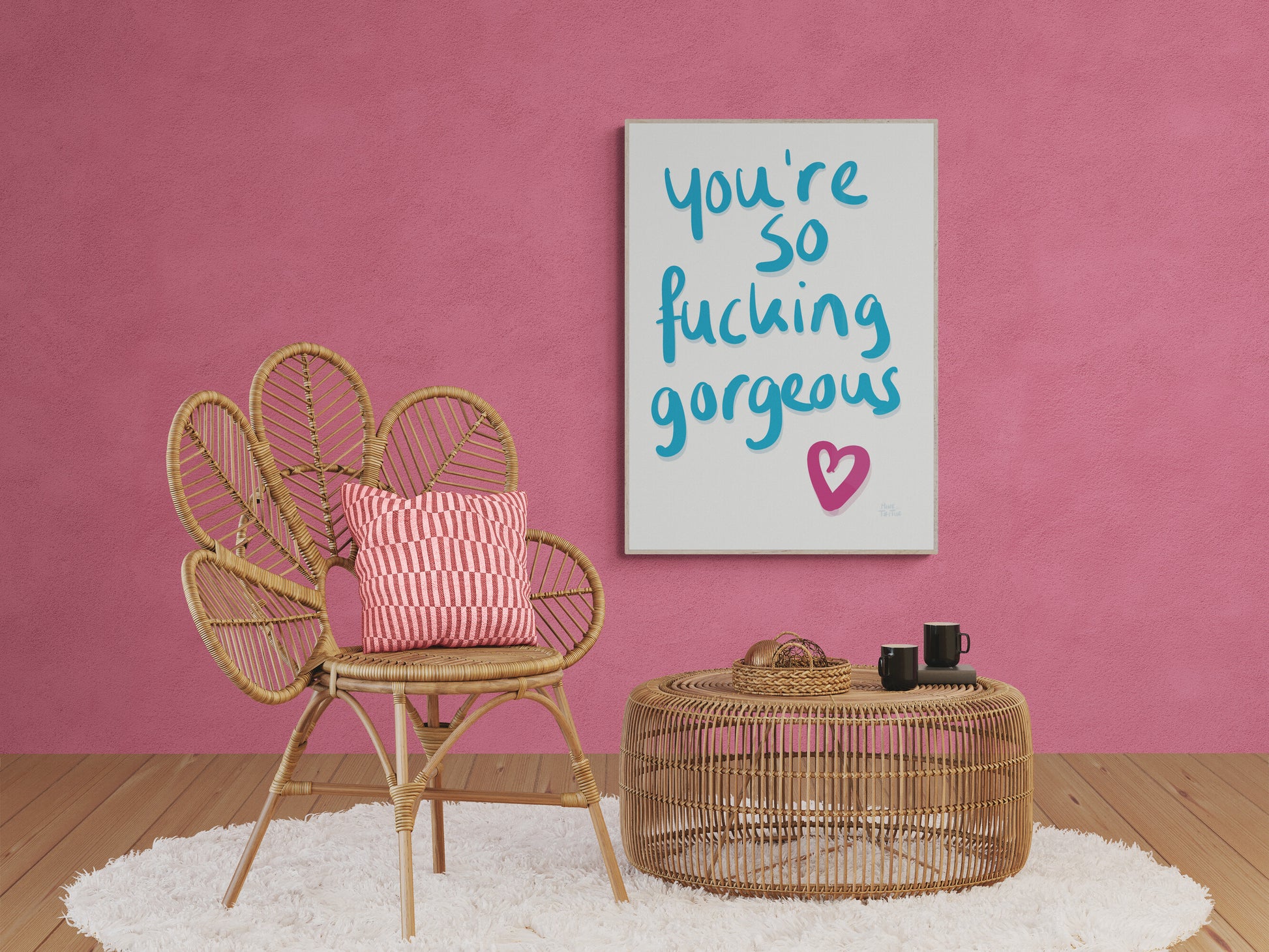 Poster with ‘You’re so fucking gorgeous’ in blue handwriting with a pink heart on a white background. Hanging on a pink wall next to wooden furniture.