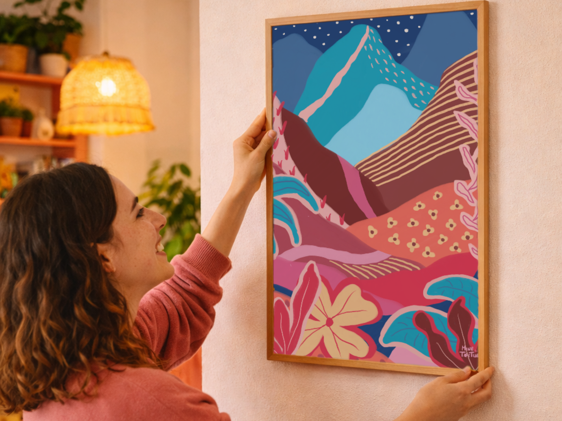 Woman hanging colourful landscape poster with mountains, sun and tropical plants on the wall. Warm, layered wall art in oak frame.